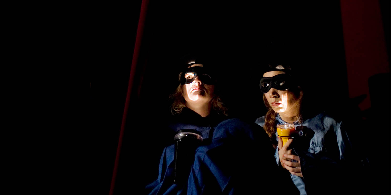 Föreställningsfoto från "Publiken", Anna Takanen och Amelie Thorén lyser med ficklampor under sina ansikten och de bär svarta masker för ögonen.